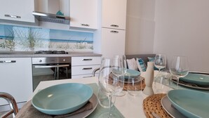 A modern kitchen with white cabinets, a gas stove, and a dining area set with blue plates and wine glasses. - Piccolo Nida 18 in Cattolica (Cattolica)