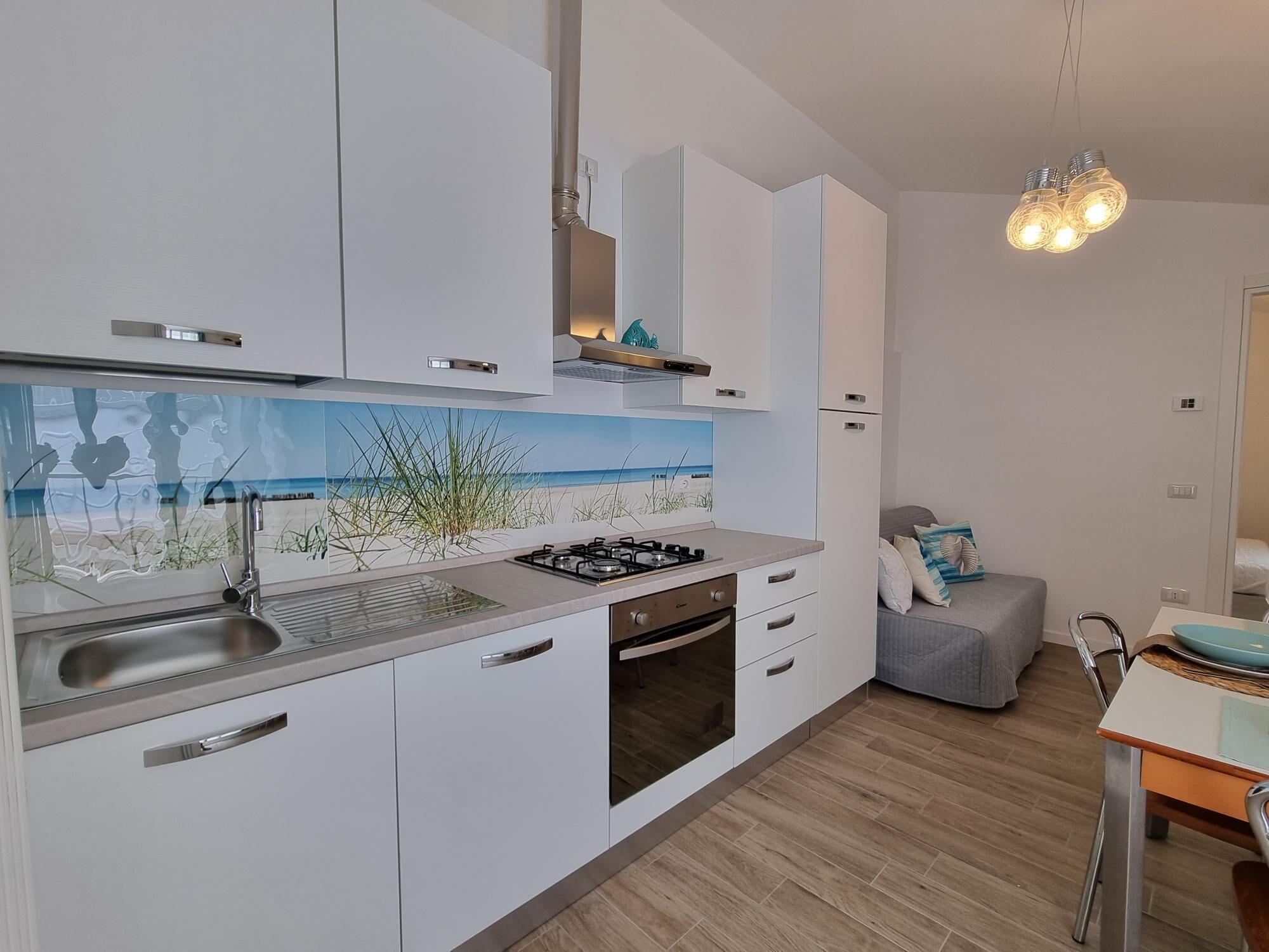Une cuisine moderne avec des armoires blanches, une cuisinière à gaz, un évier et un crédence décorée d’un motif représentant une plage. On y trouve également un coin repas avec une table et des chaises.