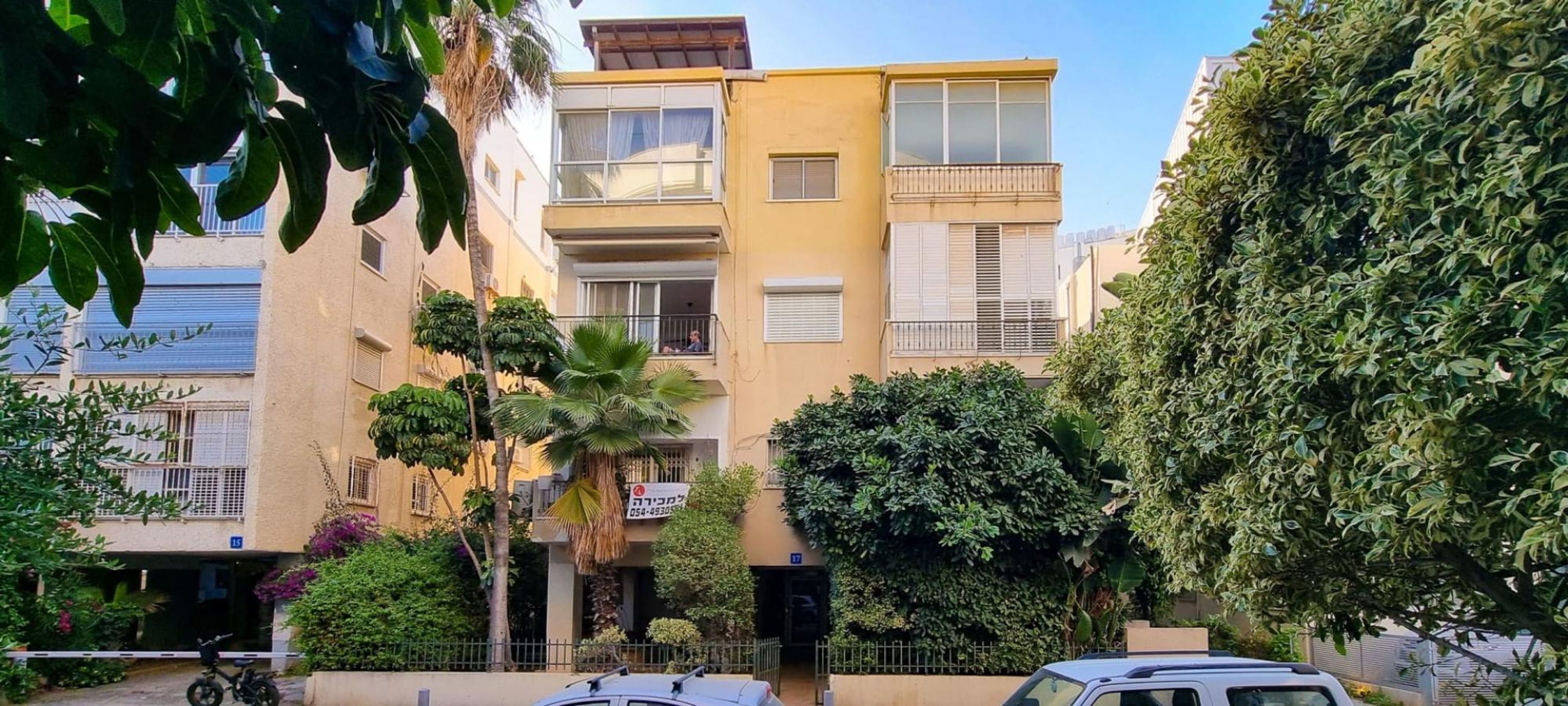 A multi-story residential building with balconies, surrounded by greenery and parked cars.