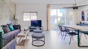 A modern living room with a brick wall, a sofa with cushions, a TV, a coffee table, an ottoman, and a dining area with chairs. - Hovevei Tsiyon Magnolia (Tel Aviv)