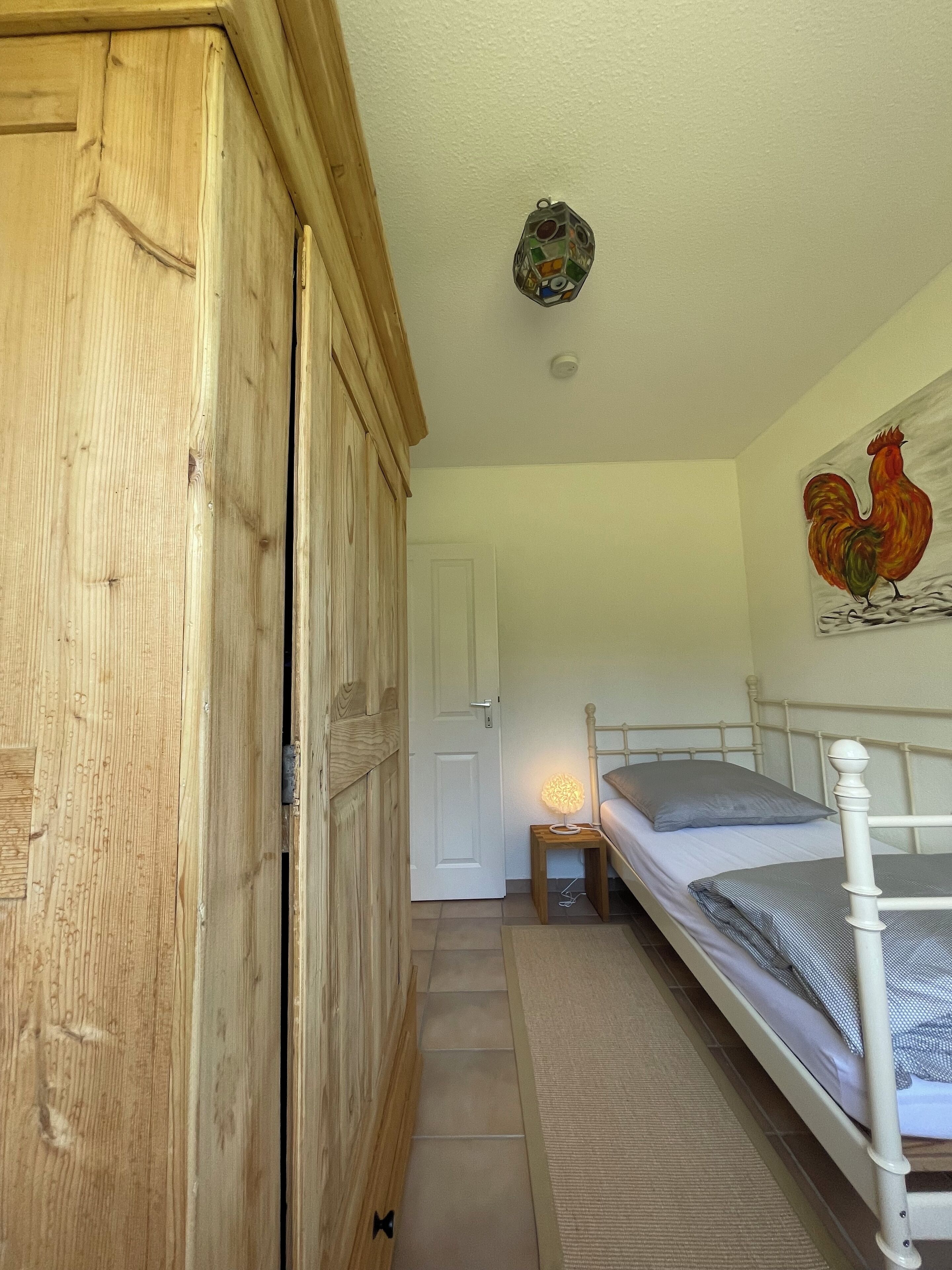 Une chambre avec une armoire en bois, un lit avec une tête de lit blanche, une petite table de chevet avec une lampe et un tableau représentant un coq, accroché au mur.