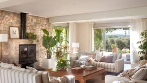 A spacious living room with a stone wall, a fireplace, and a large window offering a view of the outdoors. - Luxury villa on a private estate in Toledo w/pool and views of Sierra de Gredos. (mejorada)