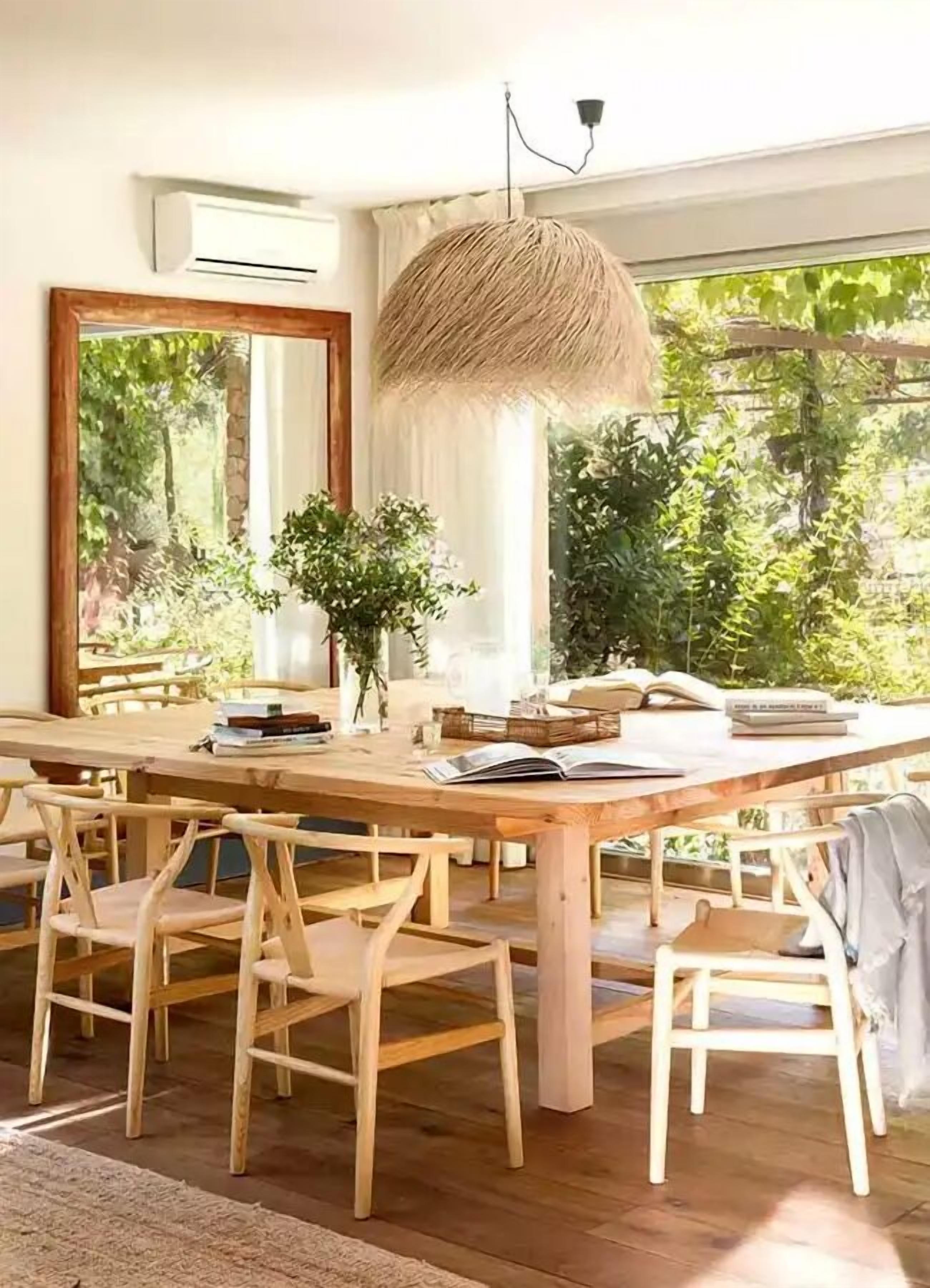 A dining area with a wooden table, six chairs, a hanging light fixture, a large mirror, and a view of greenery outside.