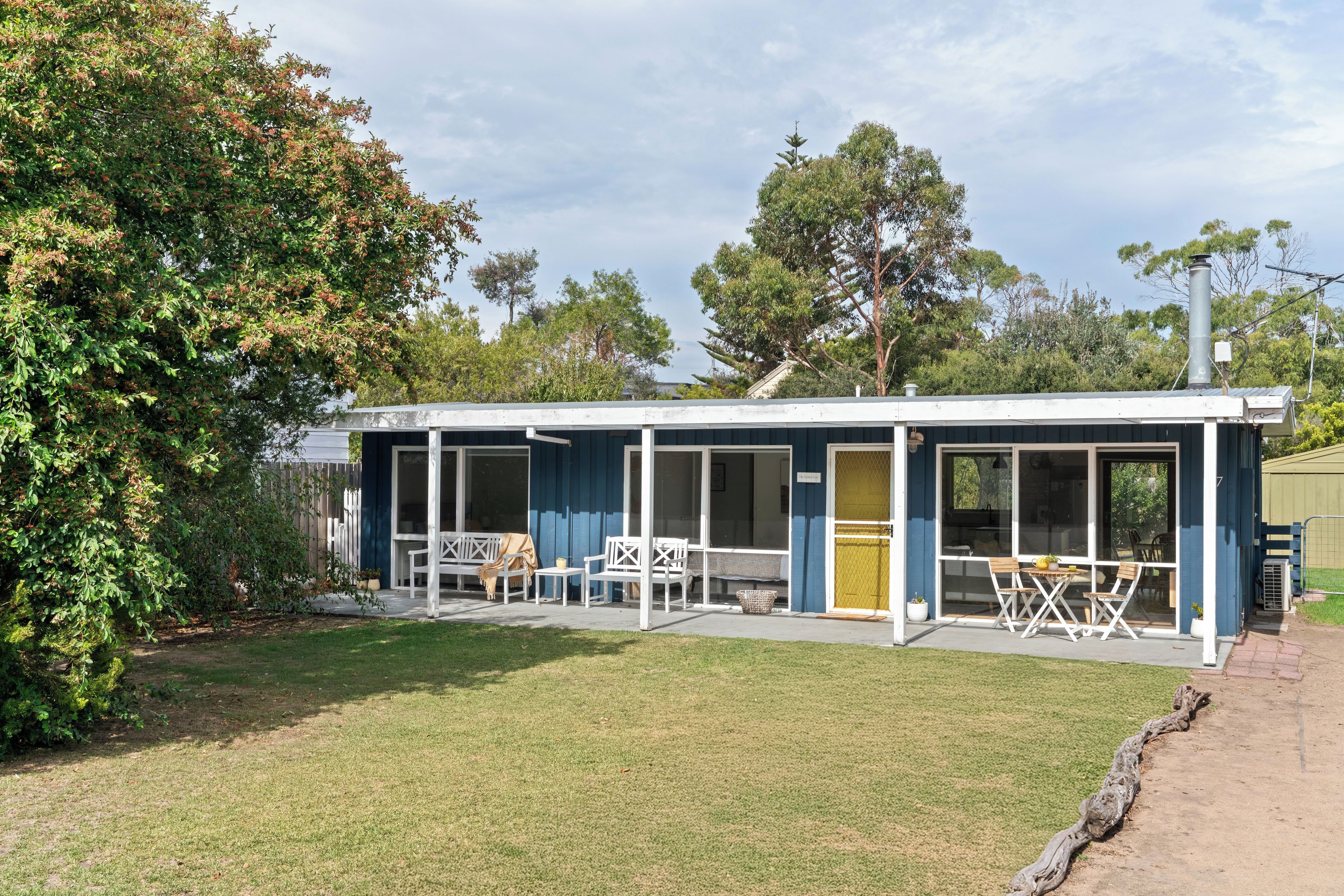 Ein eingeschossiges Gebäude mit überdachter Terrasse, einem kleinen Garten und einem großen Baum links.
