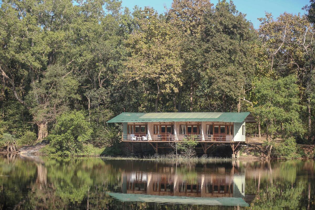 A cabin with a green roof, situated on stilts over a lake, surrounded by dense forest.