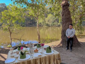 A table set outdoors with a view of a lake and trees, a person standing by the table, and a tablecloth covering the table.