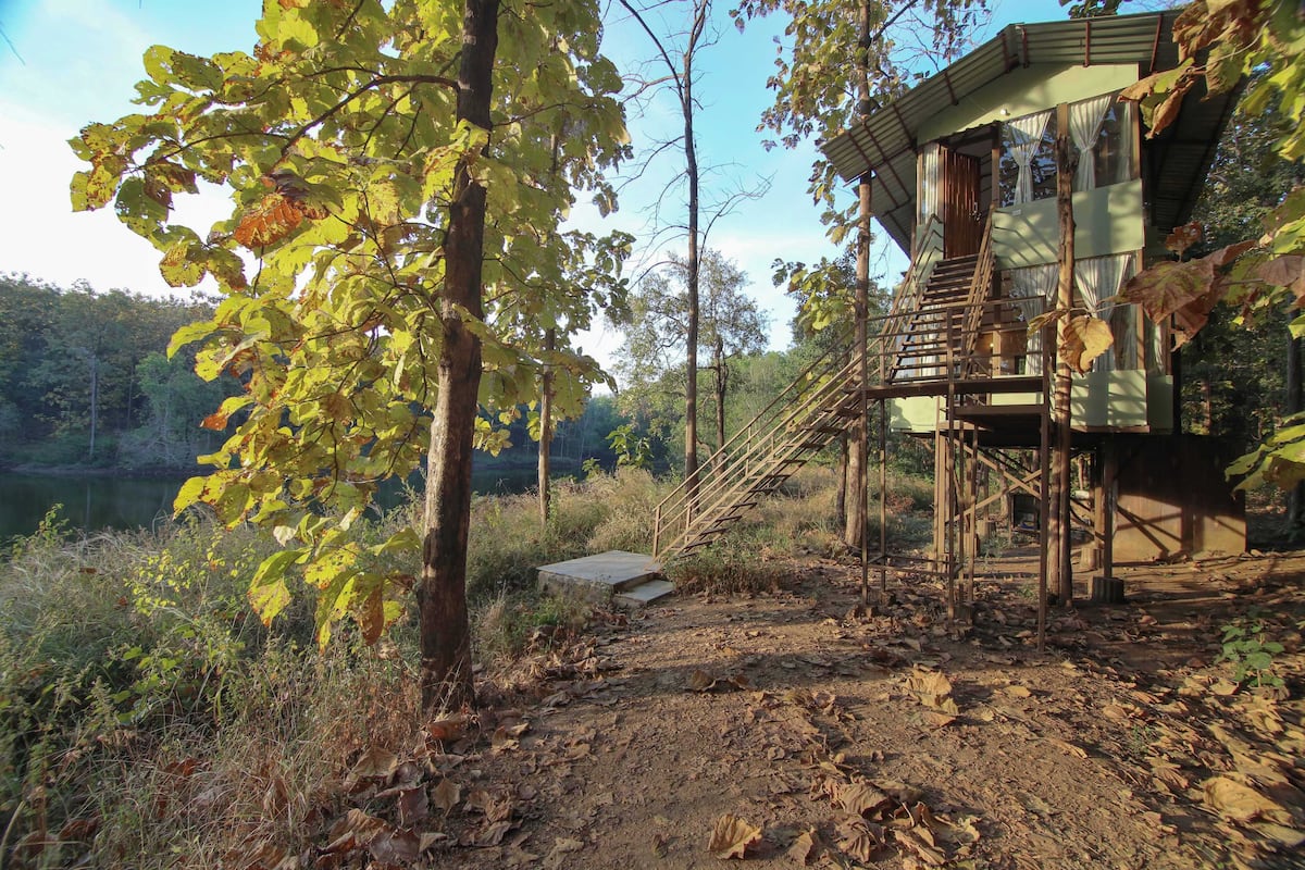 A rustic cabin with a wooden staircase leading to a loft bedroom, surrounded by trees and a dirt path.