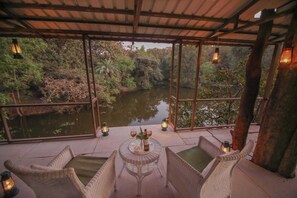 A covered outdoor seating area with wicker furniture and a table set for two, overlooking a body of water and lush greenery. - Rukhad Jungle Camp (Kurai)