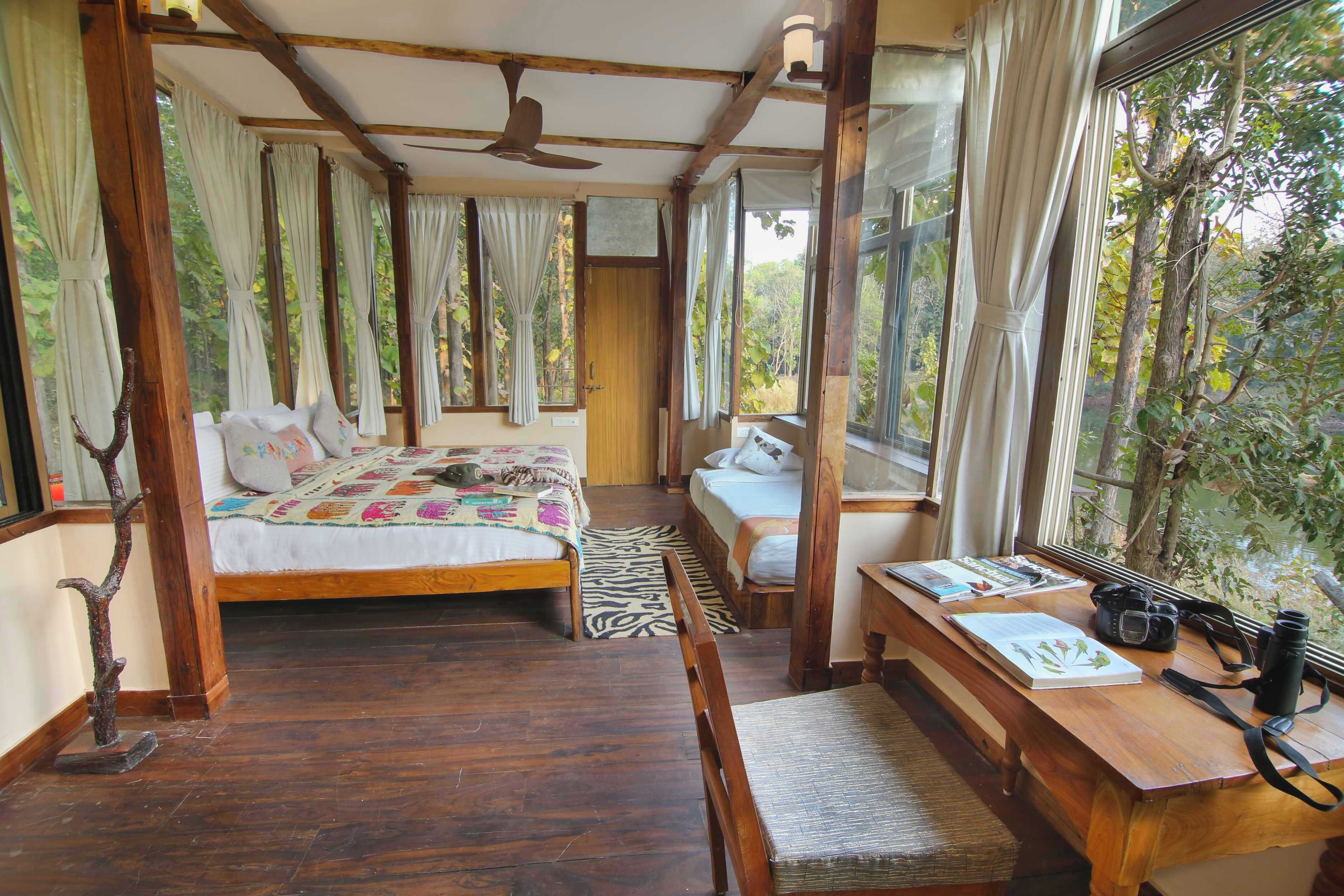 Un dormitorio con techo de madera, una cama con colcha colorida, un escritorio con cámara y botella de agua, y ventanas grandes con cortinas.