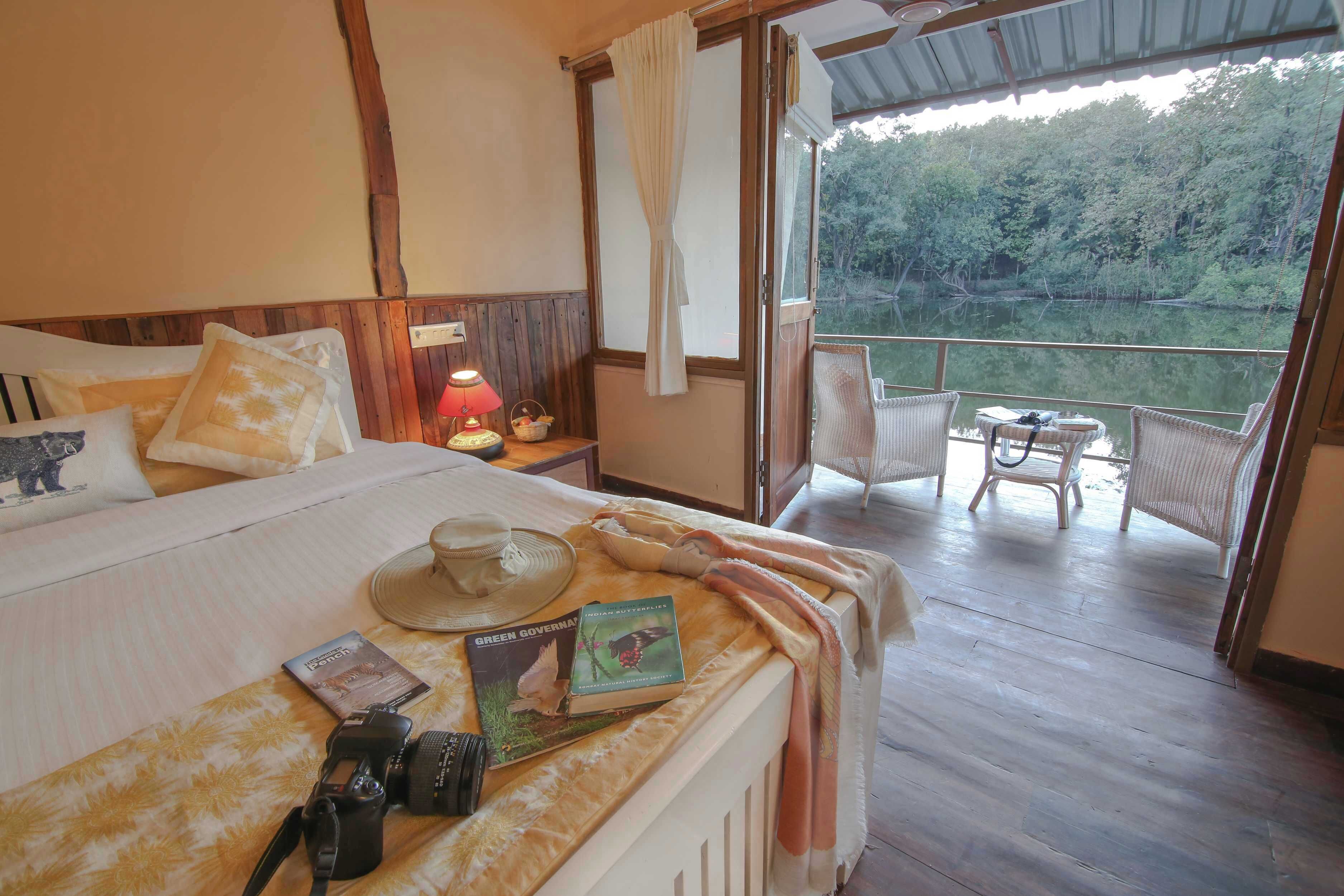 Habitación de hotel con cama, mesita de noche con lámpara, una cámara y un libro titulado 'Green Government'. Hay un balcón con sillas de mimbre y vista a un área boscosa.