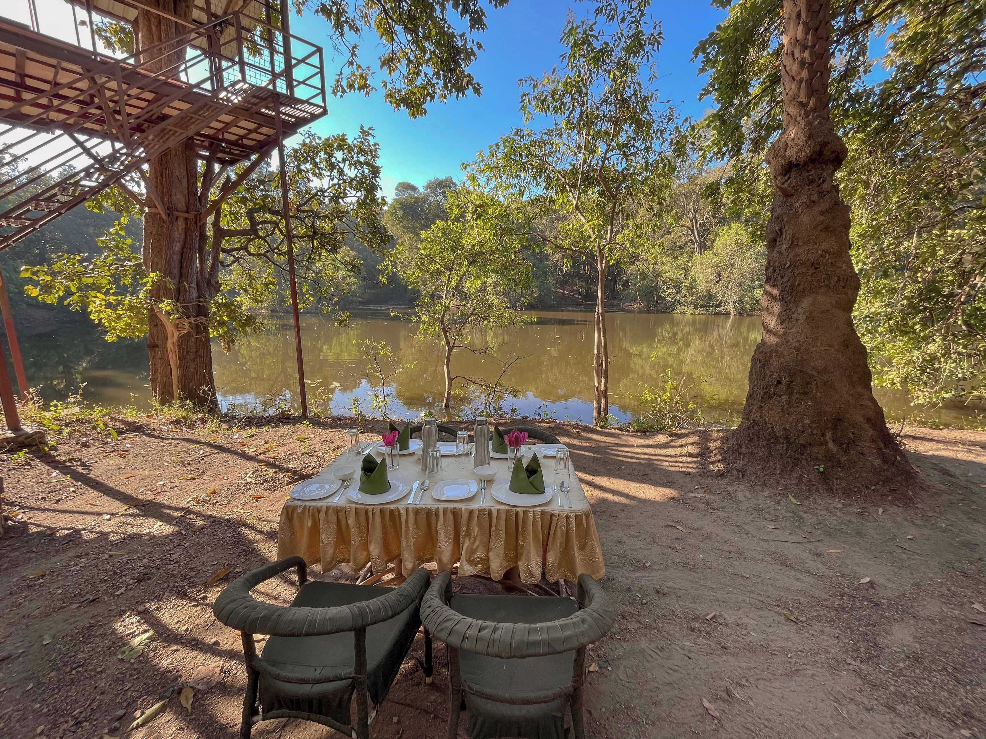 An outdoor dining setup with a table set for two, surrounded by trees and a lake view.