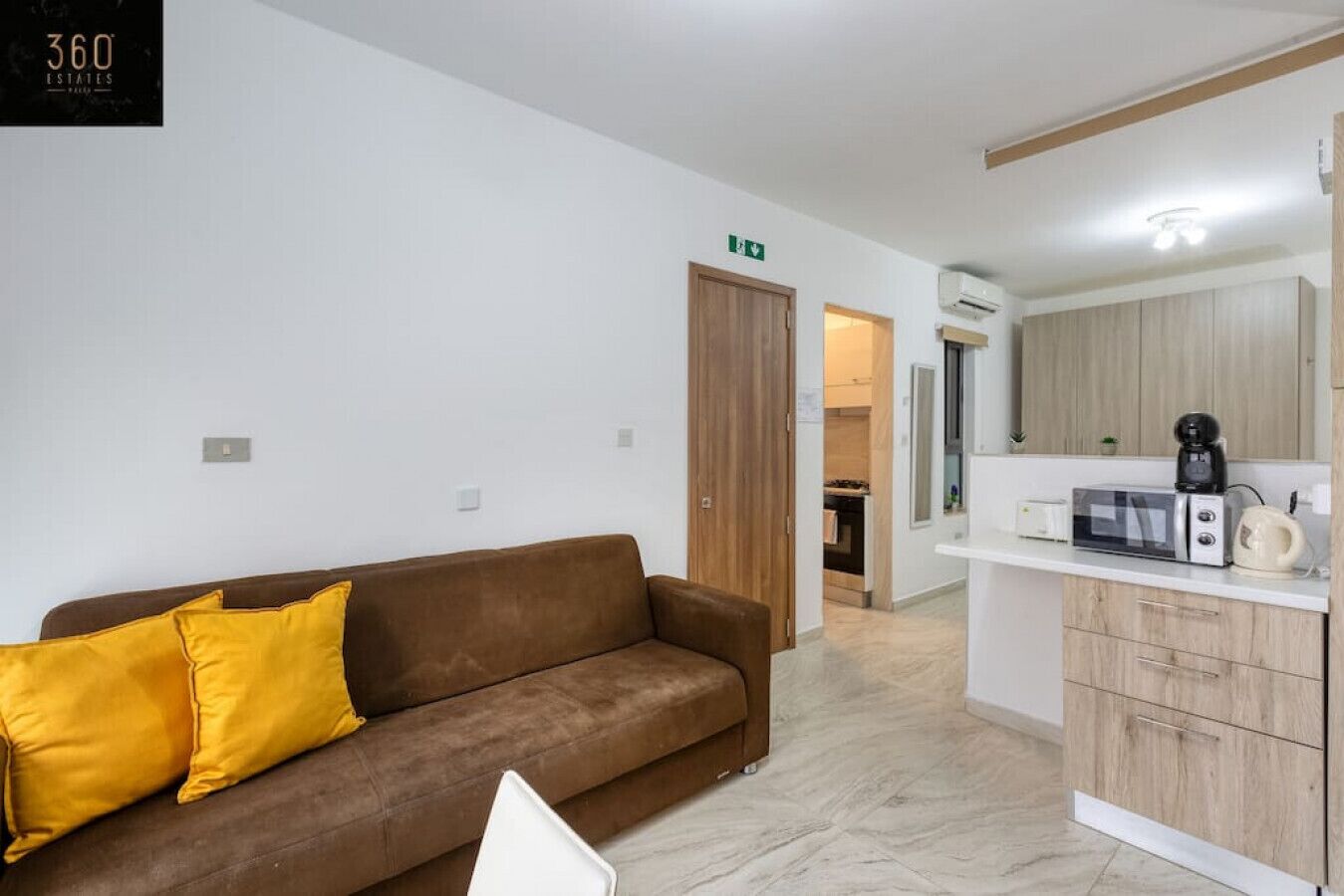 A modern living room with a brown sofa and yellow pillows, a kitchen area with a microwave and coffee maker, and a wooden door leading to another room.