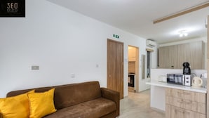 A modern living room with a brown sofa and yellow pillows, a kitchen area with a microwave and coffee maker, and a wooden door leading to another room.