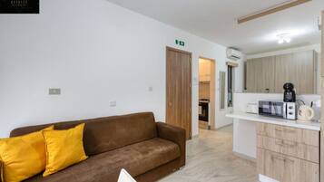 A modern living room with a brown sofa and yellow pillows, a kitchen area with a microwave and coffee maker, and a wooden door leading to another room.