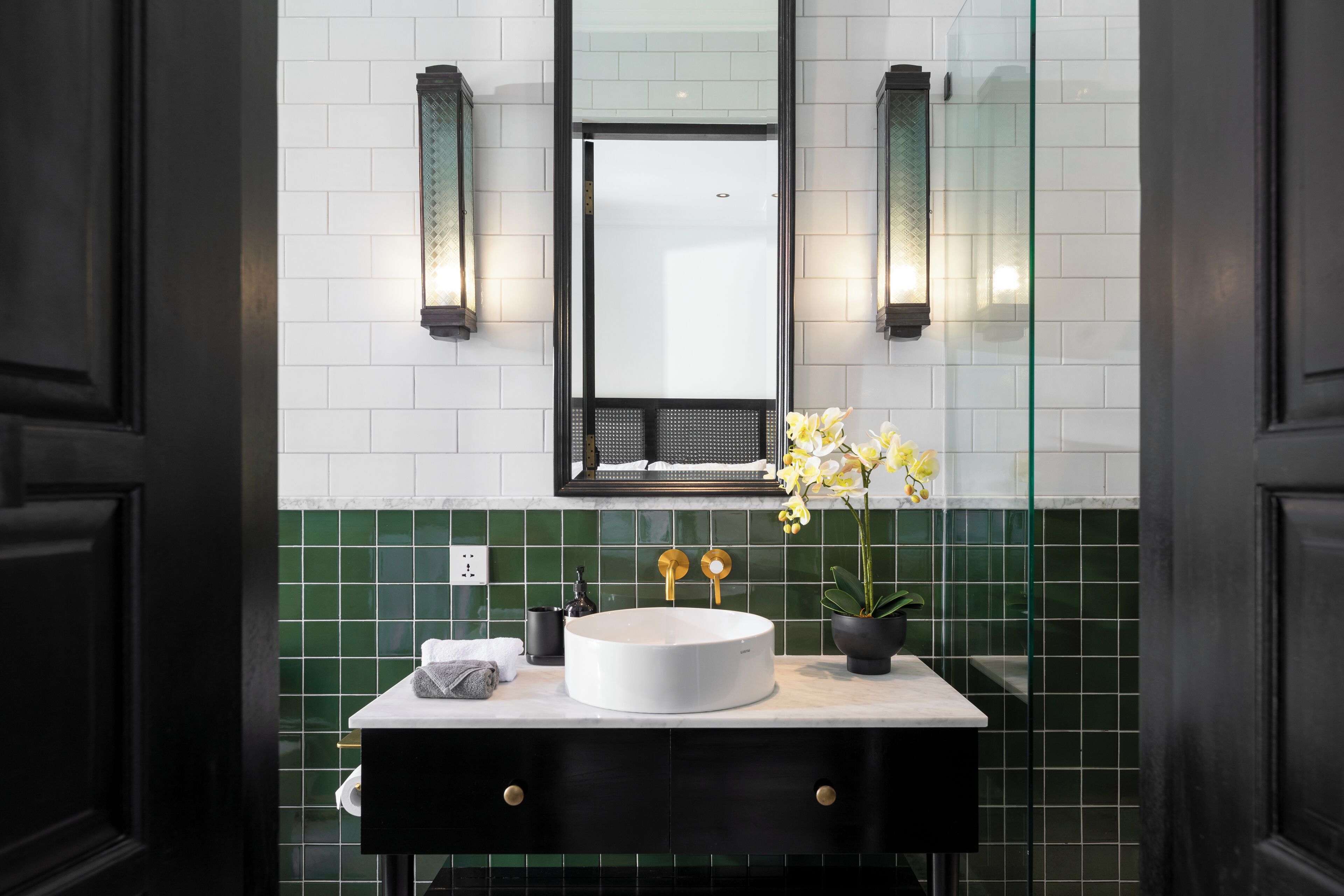 A modern bathroom with a round white sink, black vanity, green tiled backsplash, and two wall-mounted sconces.