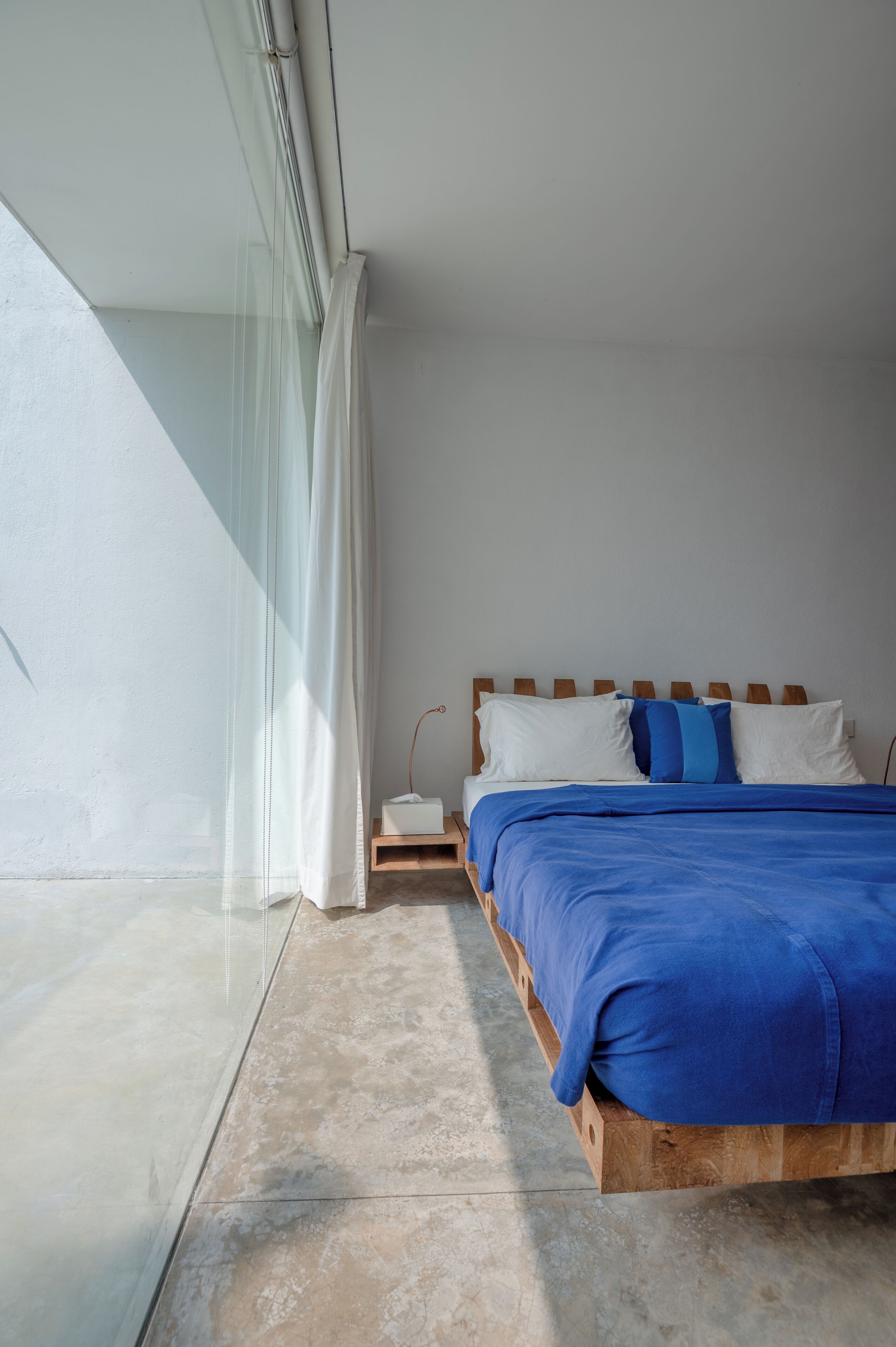 Een slaapkamer met een houten bed, beddengoed in wit en blauw, een nachtkastje en uitzicht buiten door een groot raam.