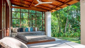 A room with a wooden ceiling, a large window with a view of lush greenery, a sofa with cushions, and a ceiling fan. - Villa Vista by The Serendipity Collection (Mirissa)