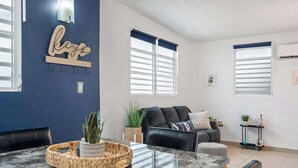 A modern living room with a dark blue accent wall, a marble countertop, a black leather sofa, and a dining area with a woven basket centerpiece. - Majestic City Home | Country Club 2 Bedroom Unit (San Juan)