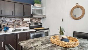 A modern kitchen with dark cabinets, a microwave, a stove, and a dining area with a marble table and woven basket. - Majestic City Home | Country Club 2 Bedroom Unit (San Juan)