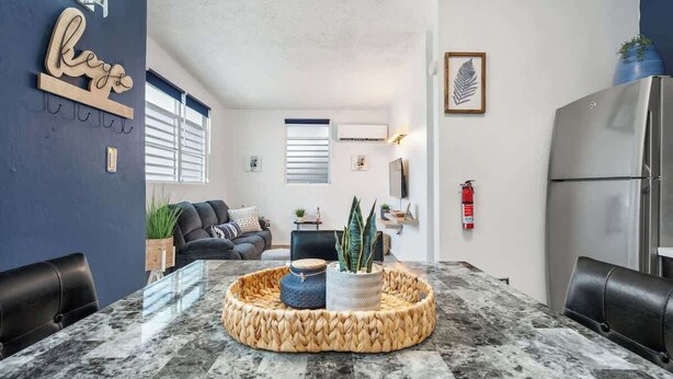 A modern living room with a glass-top dining table, a woven coffee table, a sofa, and a refrigerator. - Majestic City Home | Country Club 2 Bedroom Unit (San Juan)