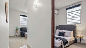 A bedroom with a bed, bedside table, and a window with blinds. - Majestic City Home | Country Club 2 Bedroom Unit (San Juan)