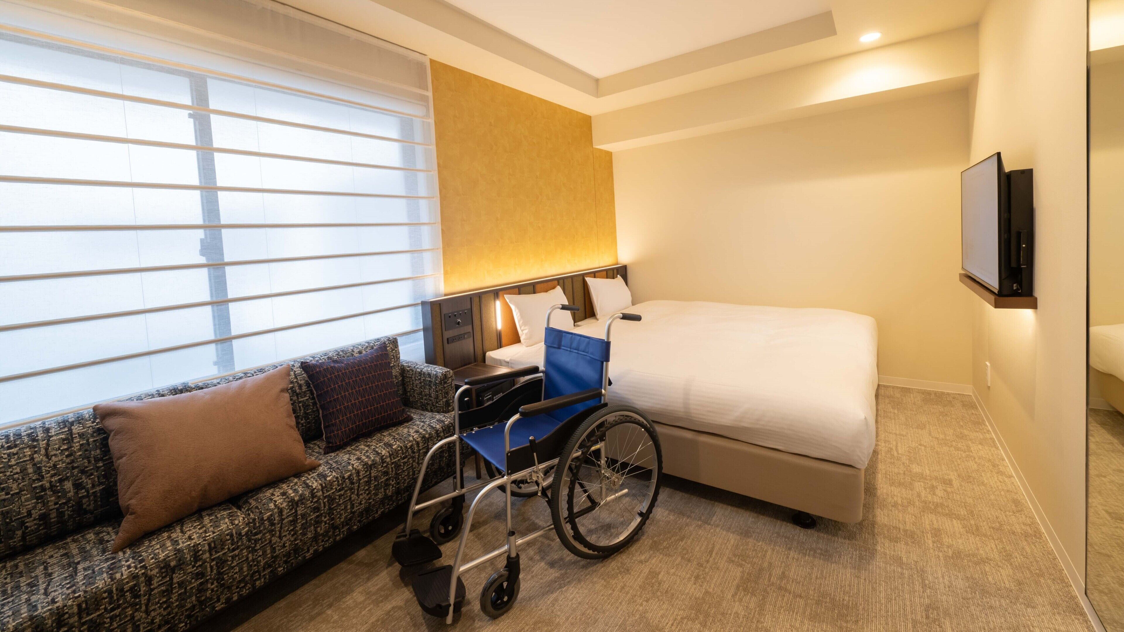 A hotel room with a sofa, wheelchair, bed, and a TV mounted on the wall.