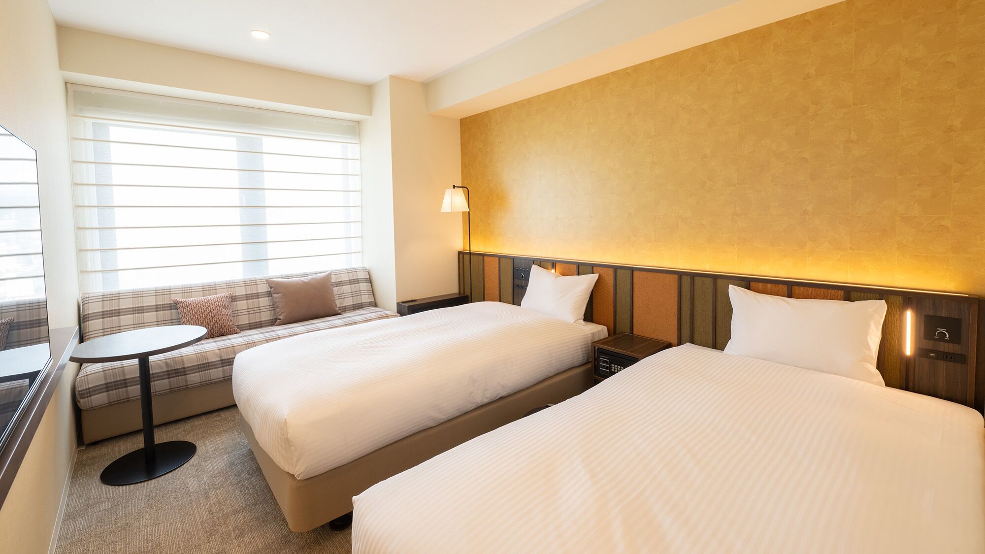 Quarto de hotel com duas camas, um sofá, uma mesa pequena e uma janela com persianas.
