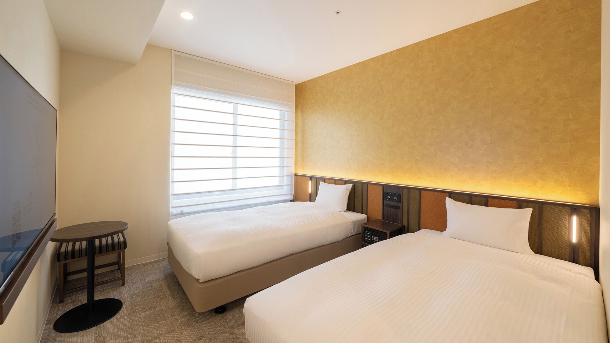 A hotel room with two beds, a TV, a small table, and a window with blinds.