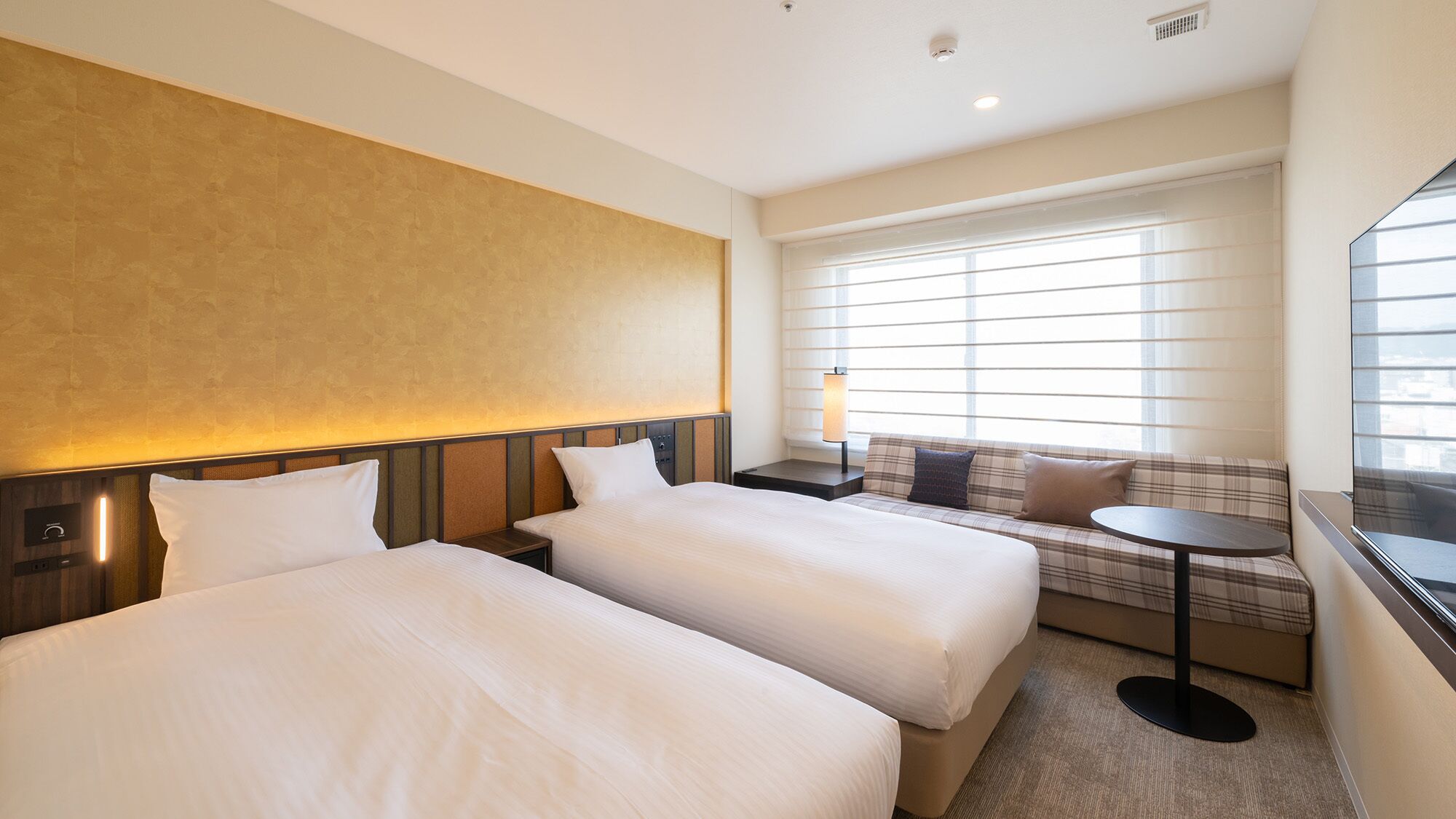Quarto de hotel com duas camas, um sofá, uma mesinha e uma janela com persianas.