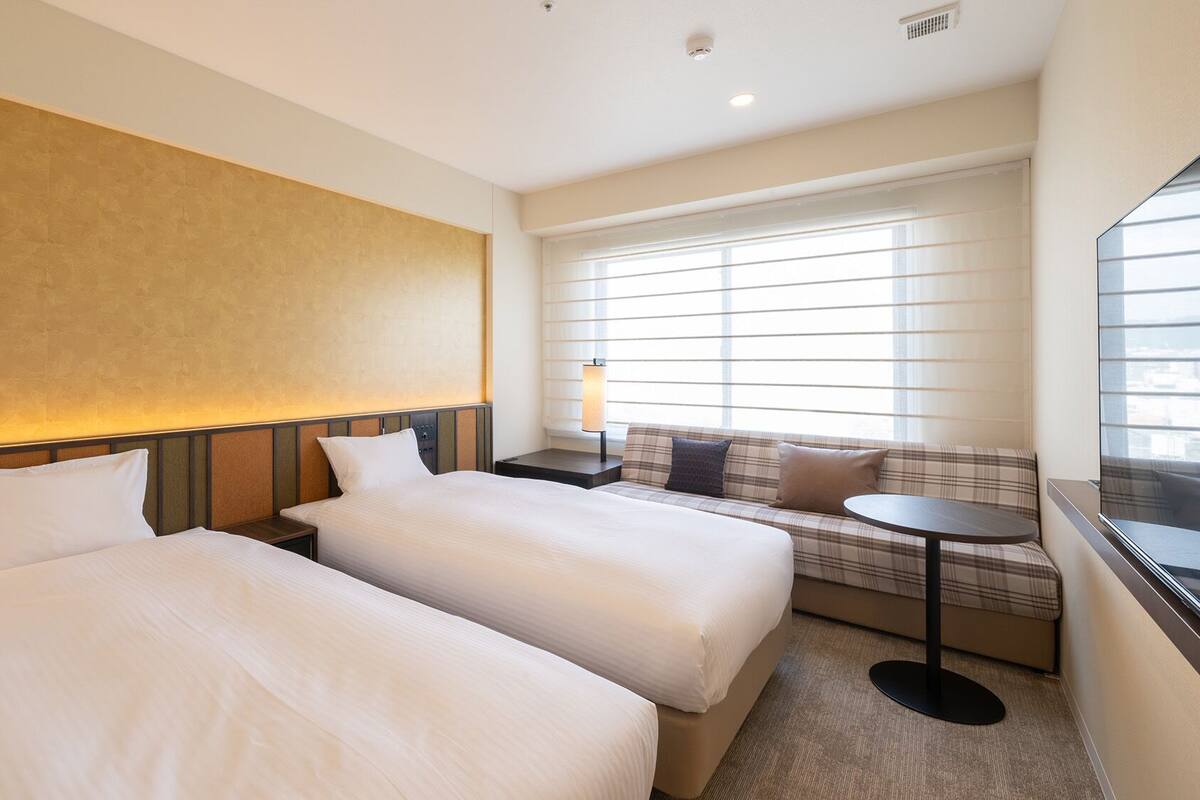 Quarto de hotel com duas camas, um sofá, uma mesinha e uma janela com persianas.