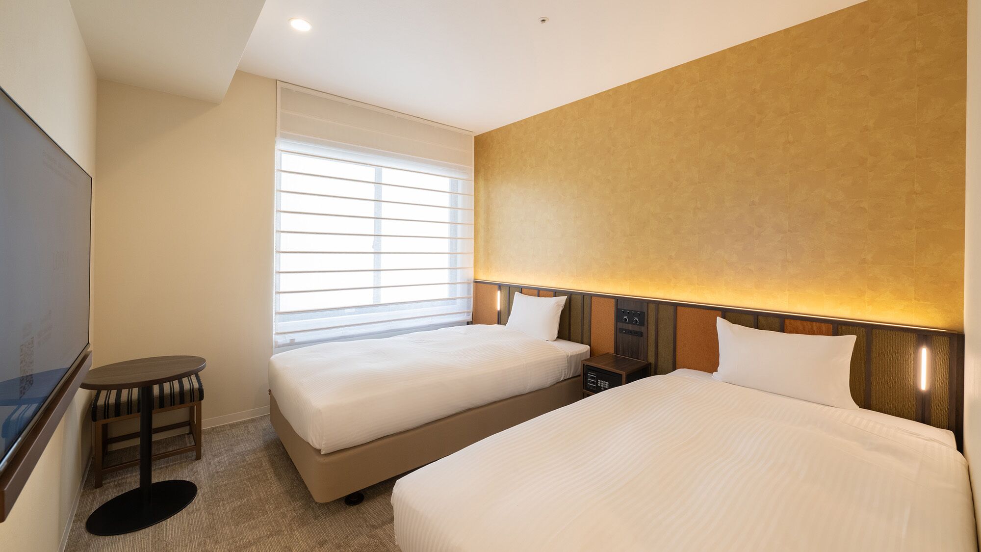 Quarto de hotel com duas camas, televisão, uma mesinha e janela com persianas.