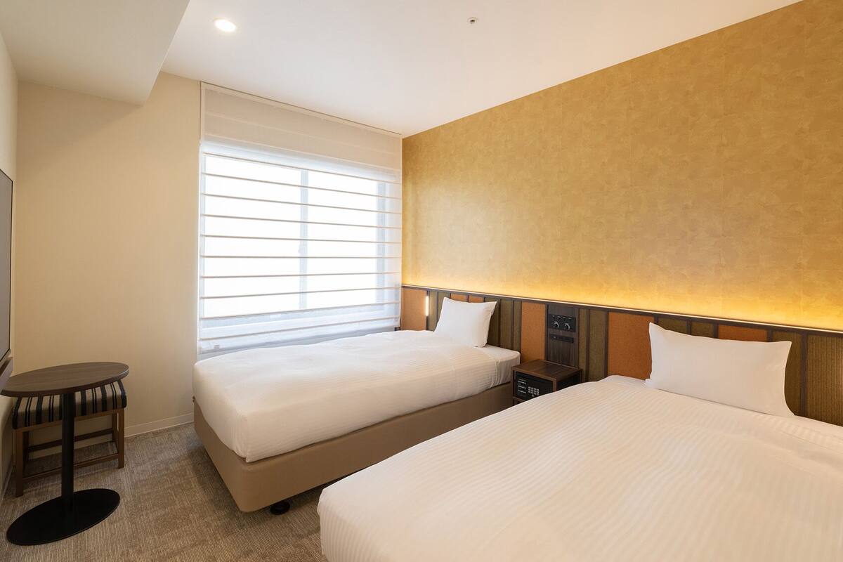 Quarto de hotel com duas camas, televisão, uma mesinha e janela com persianas.