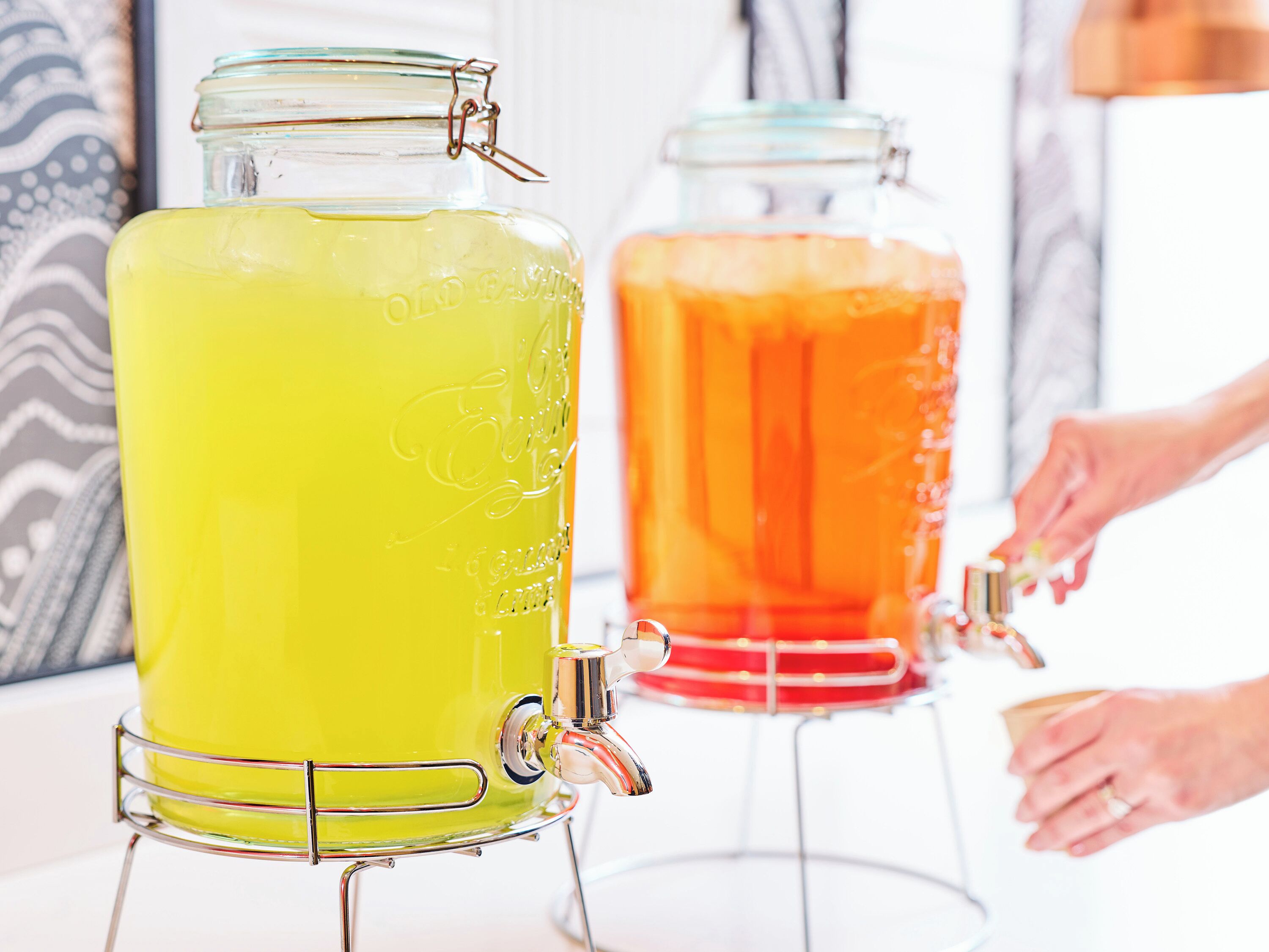 Two large glass dispensers with metal frames, one filled with a yellow liquid and the other with an orange liquid.