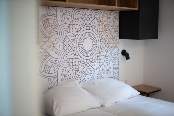 A bed with white bedding, a headboard with a geometric pattern, a small wooden nightstand, and a black wall-mounted light. - Lodge for the disabled in a holiday park (Gulpen)