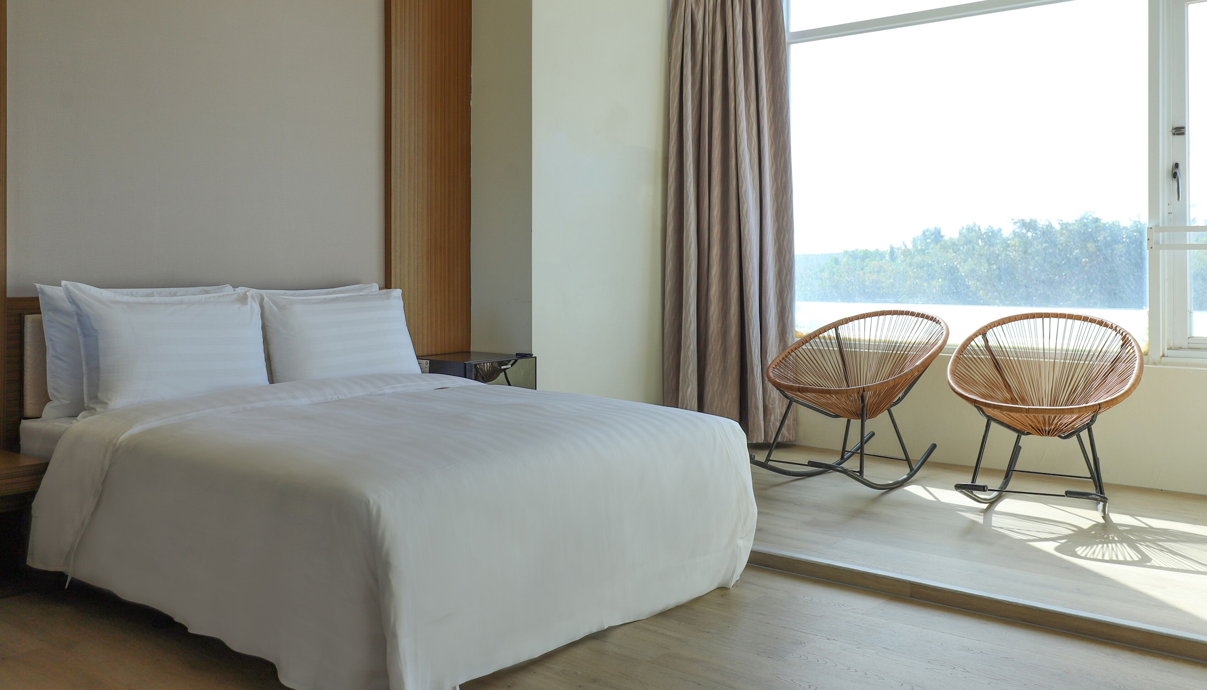 Quarto com cama grande, duas cadeiras de vime, janela com cortinas e vista para árvores.