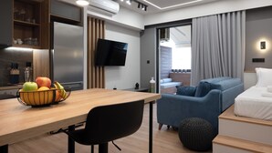 Een moderne hotelkamer met een houten eettafel, een schaal fruit, een flatscreen televisie, een bank, een bed en een groot raam met gordijnen.