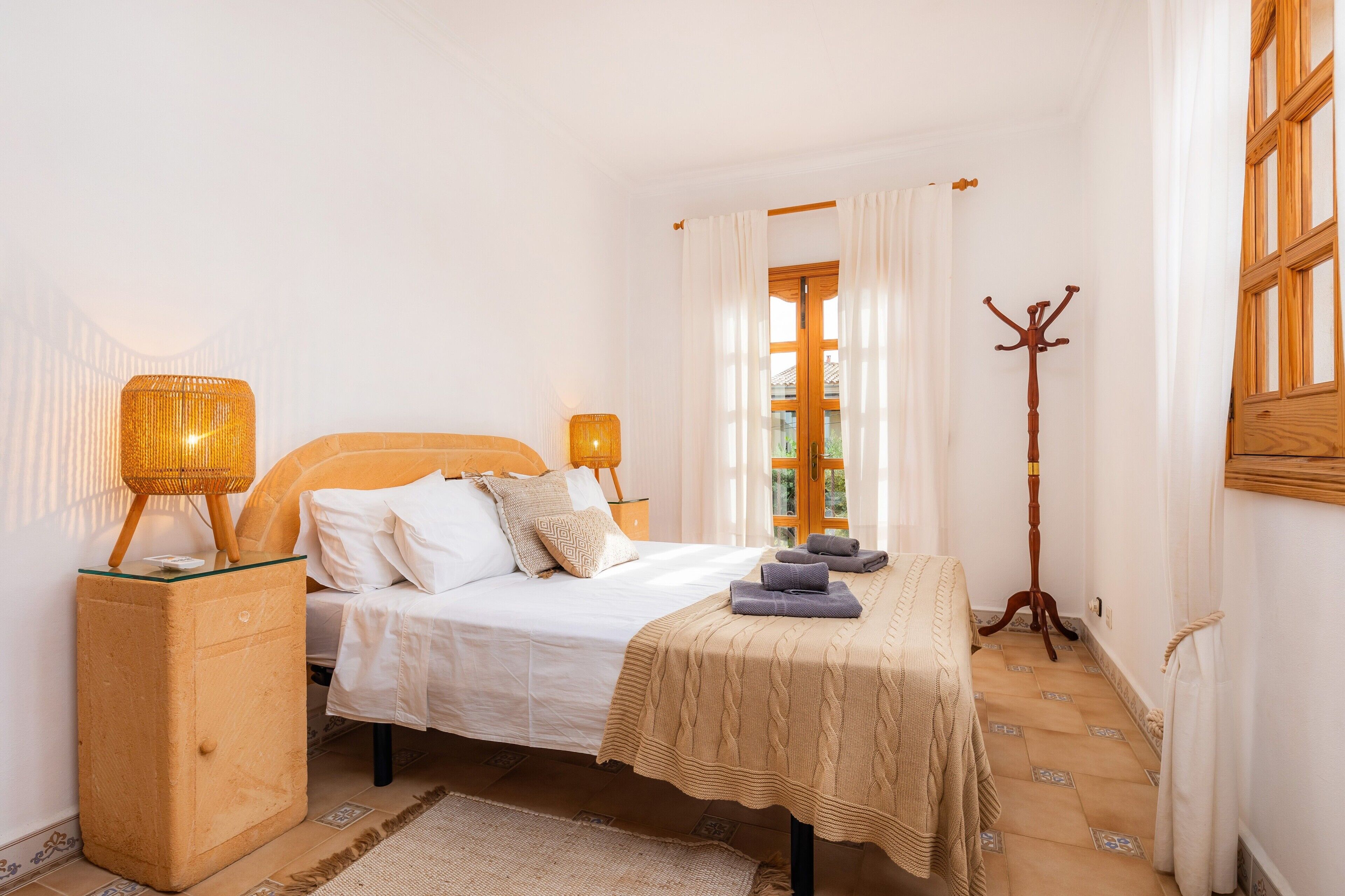 Ein Schlafzimmer mit einem Bett, Nachttischlampen, einem Holz-Kleiderständer und einem Fenster mit Vorhängen.