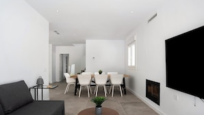 A modern living room with a sofa, a round table with a plant, a dining area with chairs, a fireplace, and a TV mounted on the wall. - Villa 'Santa Clara 21a' with Private Pool, Wi-Fi and Air Conditioning (Chiclana de la Frontera)