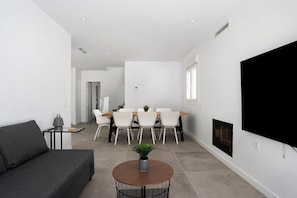 A modern living room with a sofa, a round table with a plant, a dining area with chairs, a fireplace, and a TV mounted on the wall.