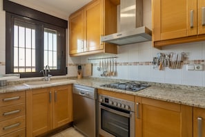 A kitchen with wooden cabinets, a dishwasher, an oven, a window with blinds, and a utensil rack. - Apartment 'Luxury Flat Beach Resort Sea View' with Shared Pool, Wi-Fi and Air Conditioning (Arenales del Sol)