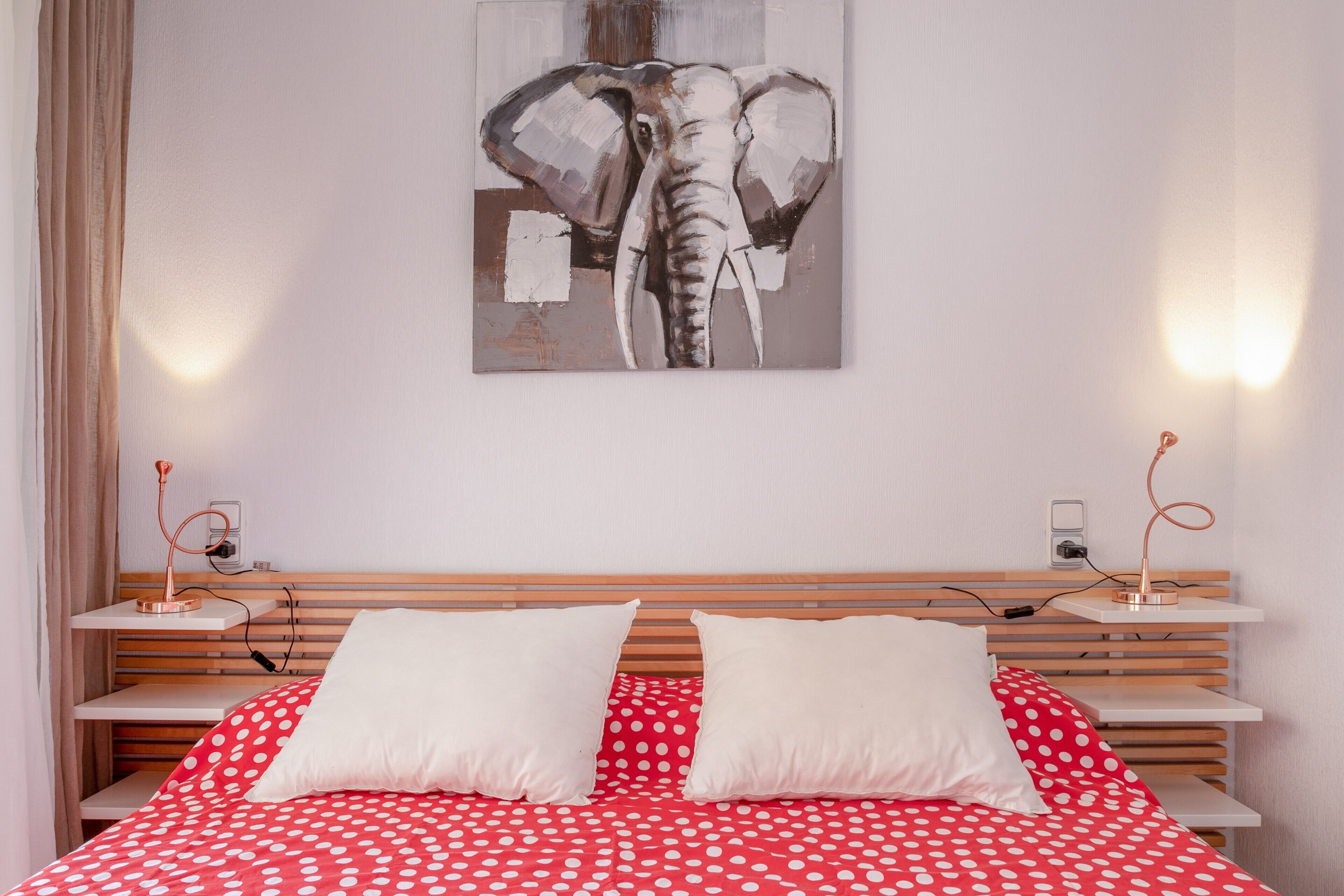 Ein Schlafzimmer mit einem Bett, Kissen, einem Holzkopfteil, einem an der Wand befestigten Elefantenbild, Nachttischlampen und einem Nachttisch.