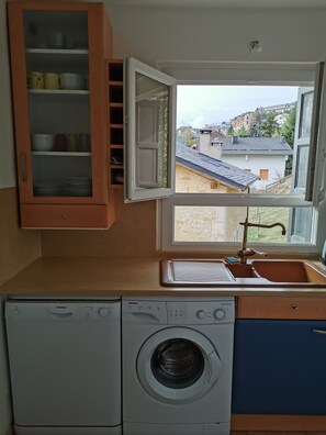 A compact kitchen with a built-in washer, a window with a view of buildings, and a wooden cabinet with dishes.