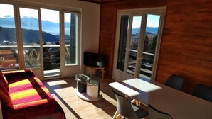 Ein Zimmer mit einer Couch, einem Esstisch mit Stühlen, einem Fernseher und großen Fenstern mit Blick auf die Berge.
