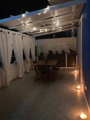 Une terrasse extérieure avec une table et des chaises, des guirlandes lumineuses et un coin cuisine agrémenté de plantes en pot.