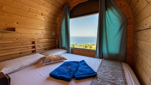 A wooden cabin room with a bed, a window with curtains, and a view of the sea. - Glamping 'Melograno' with Sea View, Wi-Fi and Air Conditioning (Tusa)