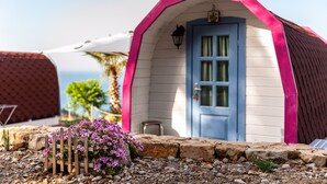 A small, single-story building with a blue door and pink trim, surrounded by a stone wall and a wooden fence with purple flowers. - Glamping 'Melograno' with Sea View, Wi-Fi and Air Conditioning (Tusa)