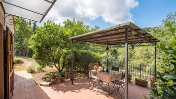 Eine überdachte Terrasse mit Tisch und Stühlen, umgeben von Blumentöpfen und Blick auf den Garten.
