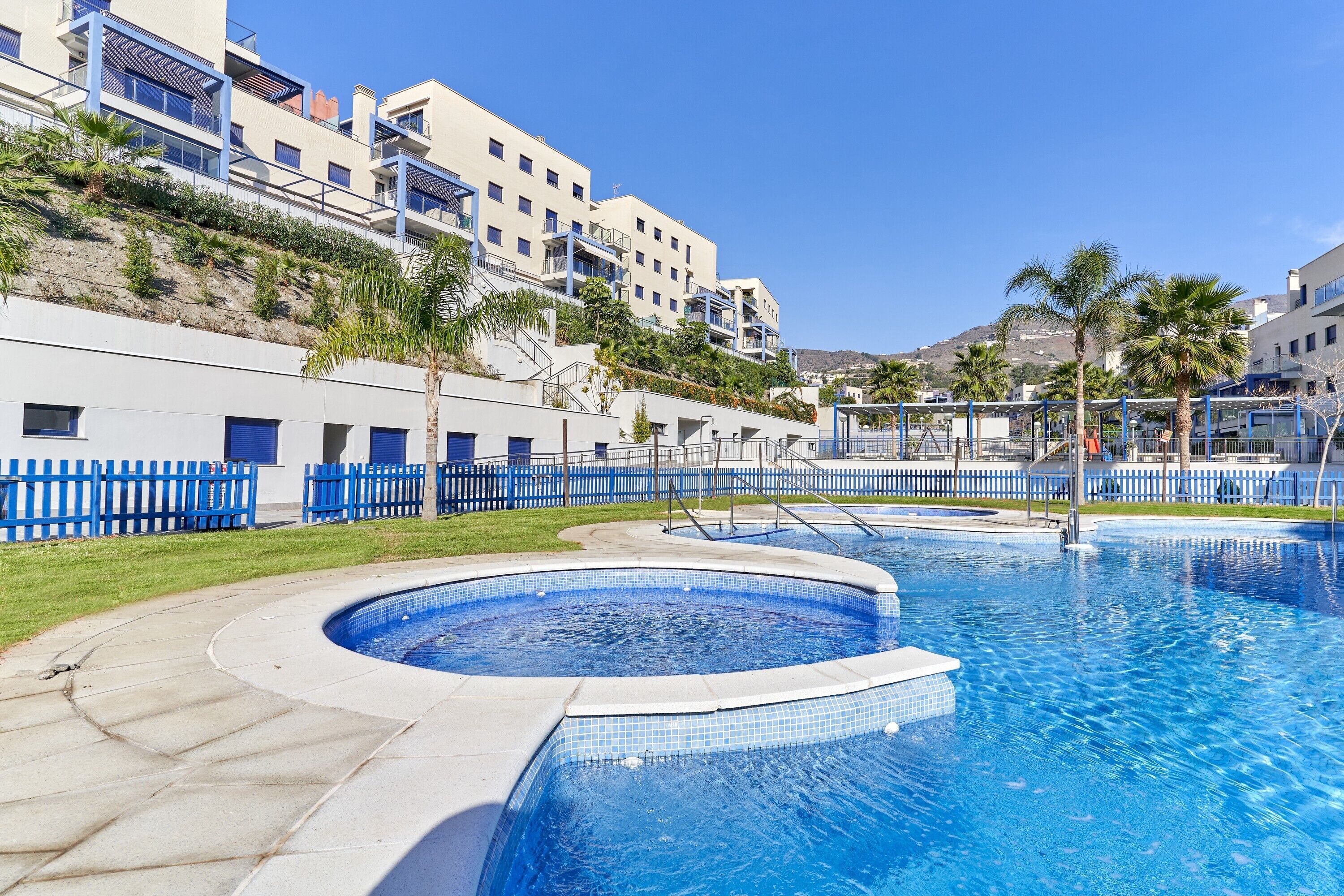 Una piscina con plataforma de azulejos, cerca delimitada por una valla azul y un edificio con balcones al fondo.