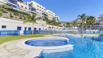 Una piscina con plataforma de azulejos, cerca delimitada por una valla azul y un edificio con balcones al fondo.