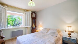 Un dormitorio con cama, mesita de noche con lámpara, reloj de madera, ventana con cortinas y radiador.